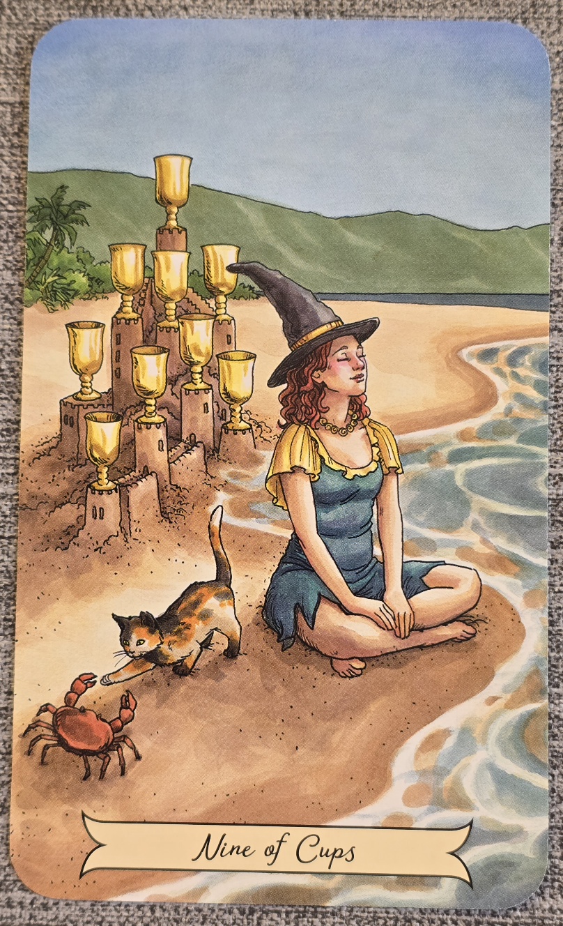 Tarotkortet Nine of Cups. På en strand sitter en rödhårig kvinna i häxhatt och färgglada kläder. Bakom henne står ett sandslott dekorerad med nio gyllene bägare. En svart katt leker med en liten röd krabba i förgrunden. I bakgrunden syns havet, en strand och gröna kullar under en ljusblå himmel.