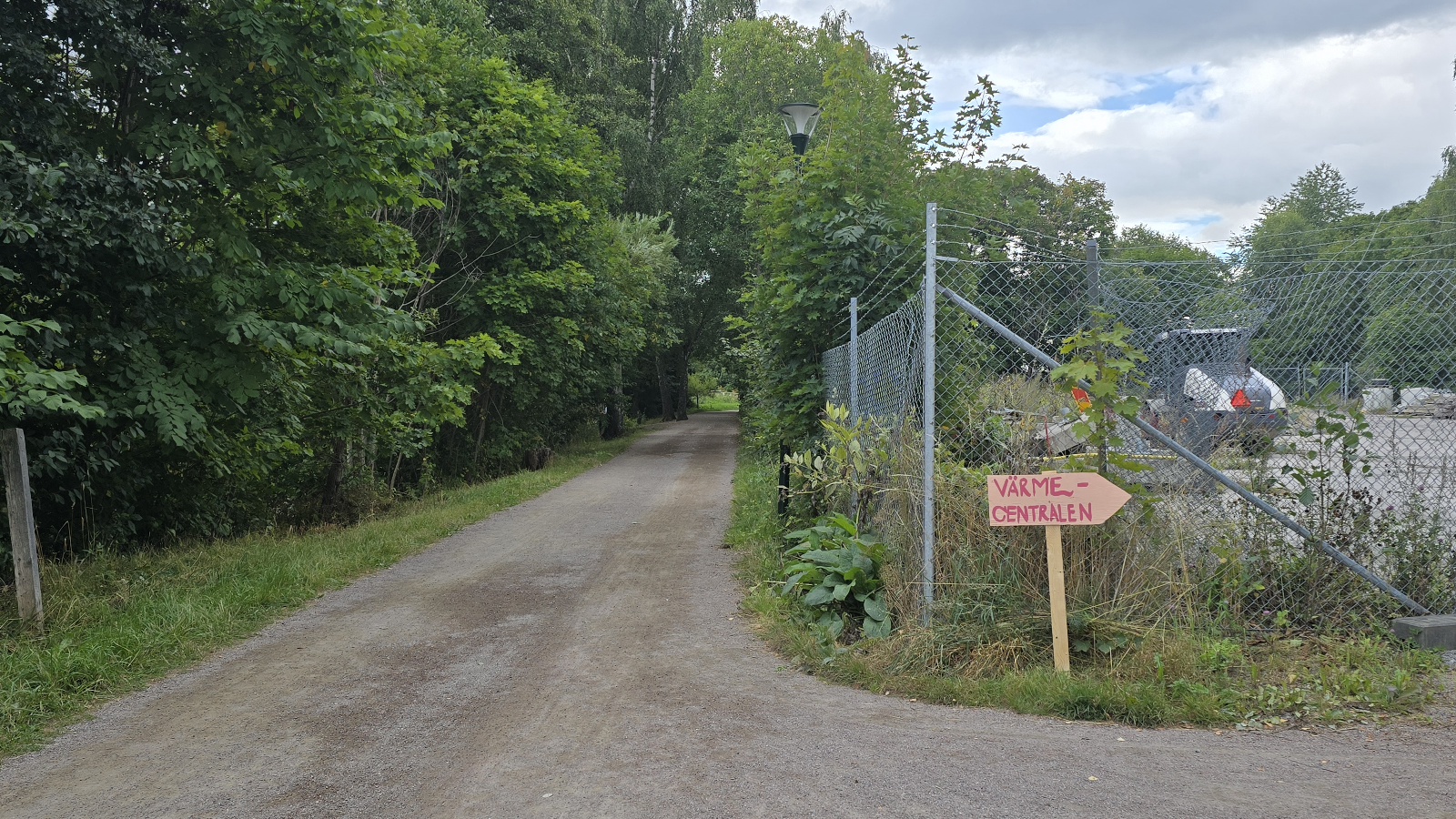En grusväg går genom ett lummigt grönområde. Till höger finns ett inhägnat område med teknisk utrustning och en pilskylt märkt Värmecentralen som pekar åt höger. Upp mot vägen syns träd och en delvis molnig himmel.