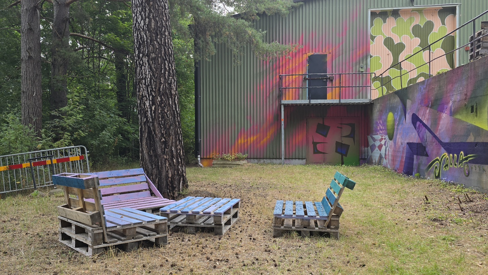 En skogsdunge omger en öppen grus- och gräsbevuxen plats med flera hemmagjorda sittplatser av träpallar i blå och lila färger. I bakgrunden står en grön korrugerad byggnad med färgstark graffiti, bland annat hjärtan och abstrakta motiv. En målad mur syns till höger.