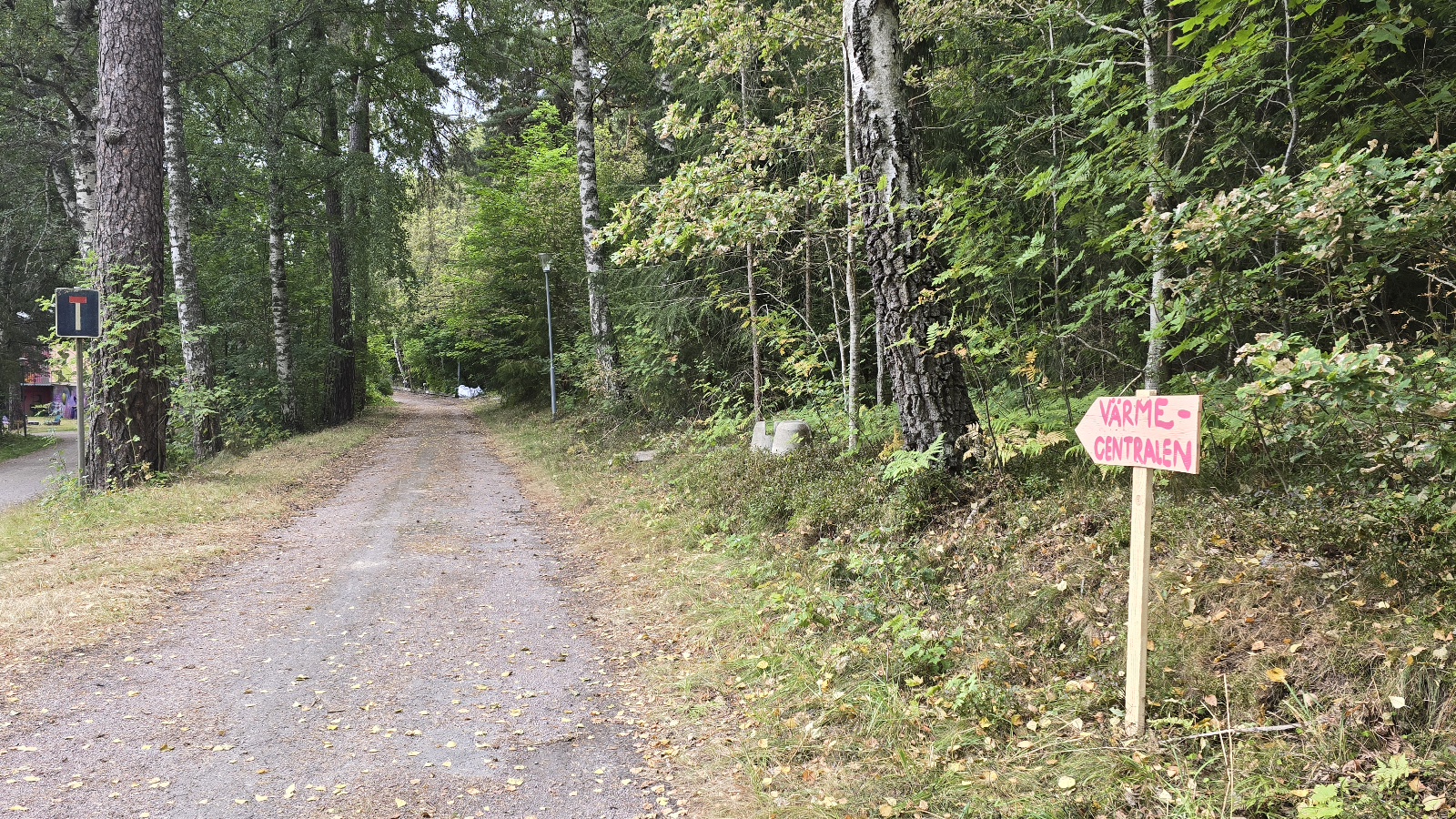 En smal grusväg går genom ett skogsområde. Till höger står en handmålad skylt med texten Värmecentralen och en pil som pekar åt vänster. Träd och tät vegetation omger vägen.