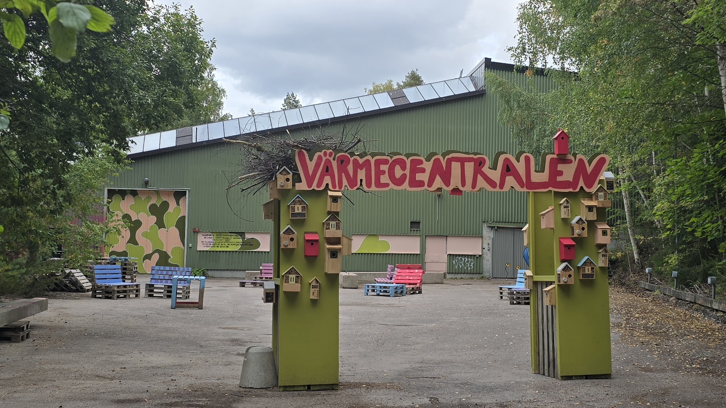 En grön portal med texten VÄRMECENTRALEN högst upp leder in på en grusad gårdsplan framför en låg, grön byggnad. Portalens två pelare är dekorerade med små fågelholkar och färgglada detaljer. Runt platsen står träd, och i bakgrunden syns utemöbler samt en färgglad väggmålning.