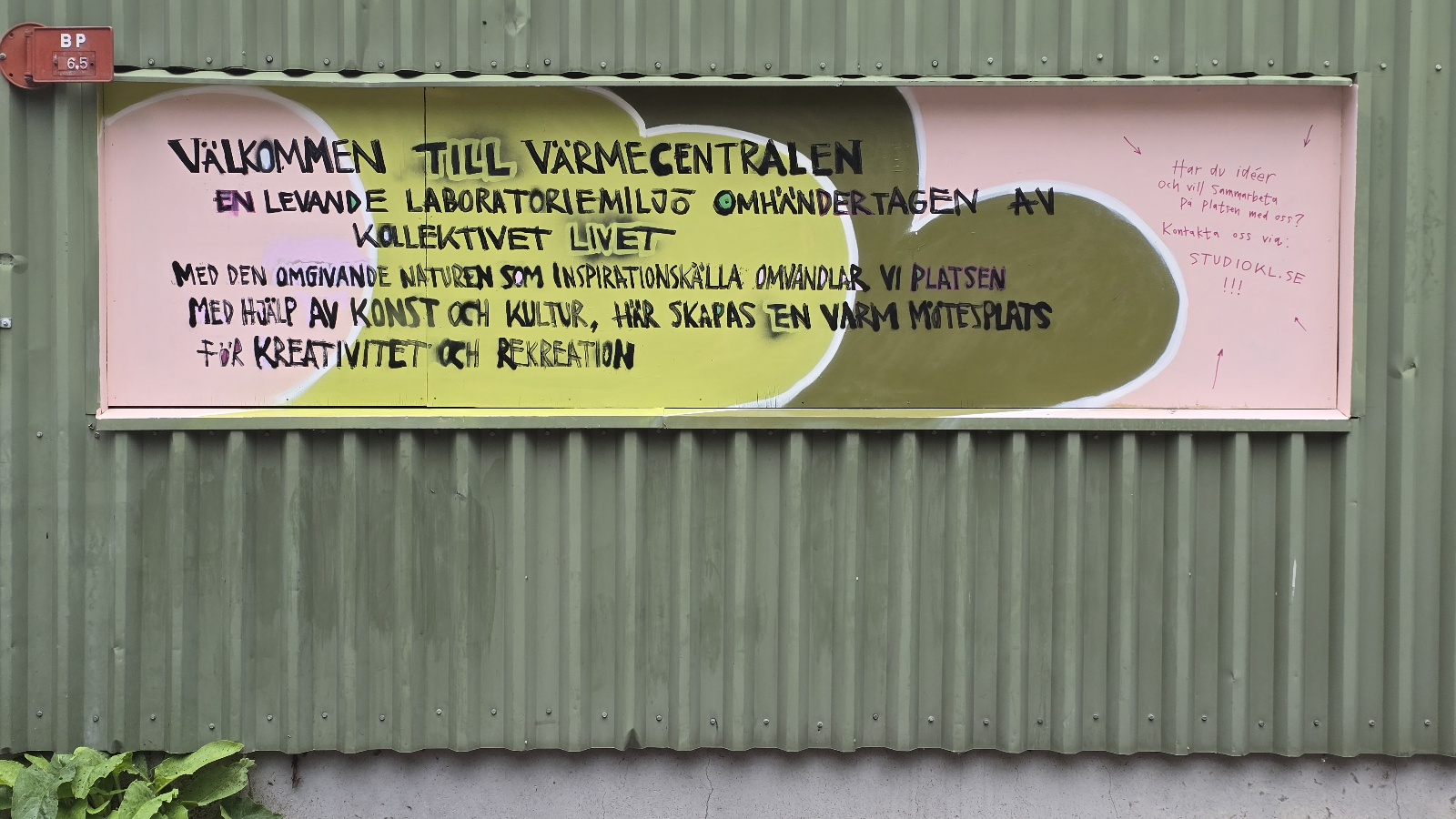 En rosa informationsskylt på en grön korrugerad plåtvägg. Överst står det: Välkommen till Värmecentralen. Under rubriken finns handskriven text om platsen. Skylten har färgfält i gult, vitt och mörkgrönt samt små handskrivna anteckningar på högra sidan. Vegetation syns längst ned i bilden.
