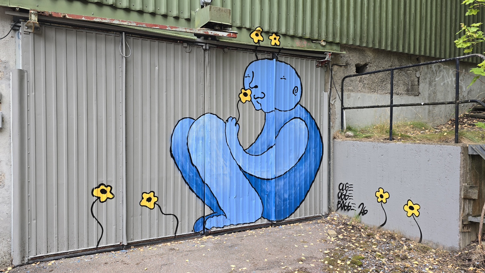 En graffitimålning på en grå korrugerad plåtdörr visar en blå, stiliserad figur som sitter hopkrupen och håller om sina knän. Figuren har ett runt huvud med en enkel ansiktsprofil. Runt figuren finns små gula blommor med svarta, böjda stjälkar. Till höger syns en låg grå betongvägg med svart graffiti, och ovanför löper en grön korrugerad vägg.