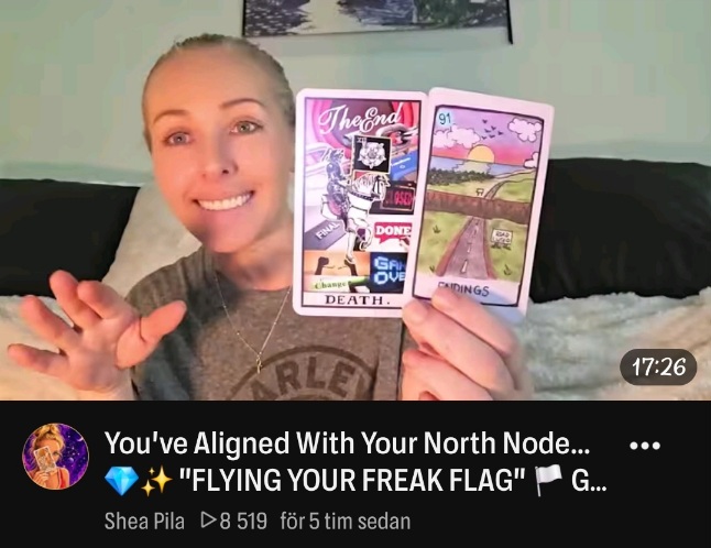 En person ler mot kameran och håller upp två tarotkort bredvid varandra. Personen sitter i en soffa. Bilden är en skärmdump från en video med texten: You’ve Aligned With Your North Node… FLYING YOUR FREAK FLAG